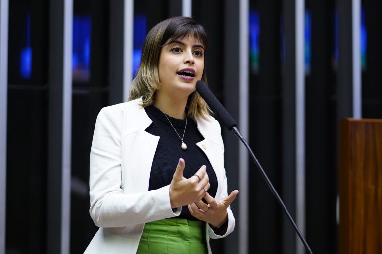 Deputada Tabata Amaral fala ao microfone. Ela é branca, tem o cabelo escuro, usa uma blusa preta, blazer branco e saia verde
