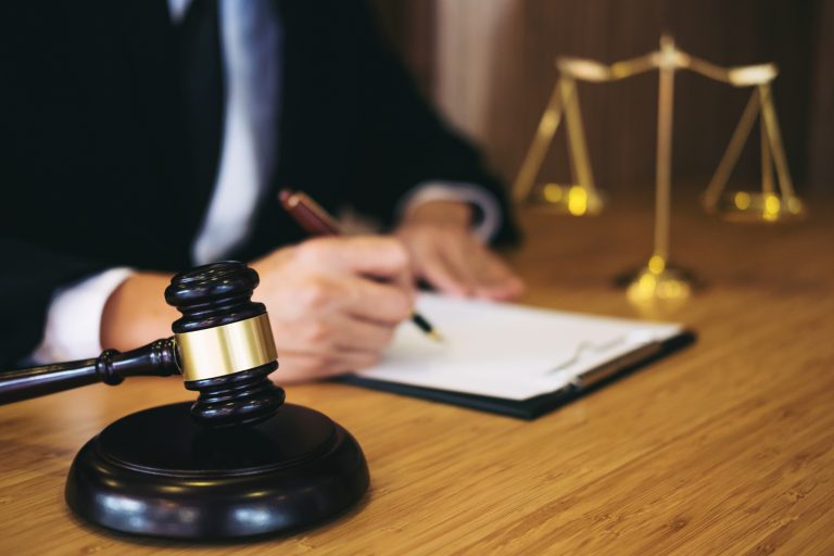 Um homem assina um papel, ao lado há um martelo de juiz e uma balança representando a justiça