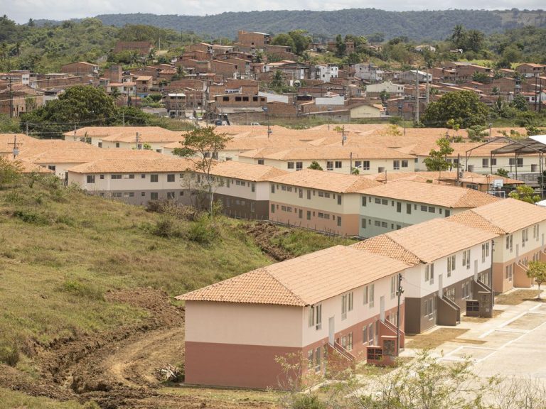 Vista de complexo de apartamentos. Prédios de dois andares pintados em cores claras. Ao fundo há um morro com vegetação e o dia está ensolarado