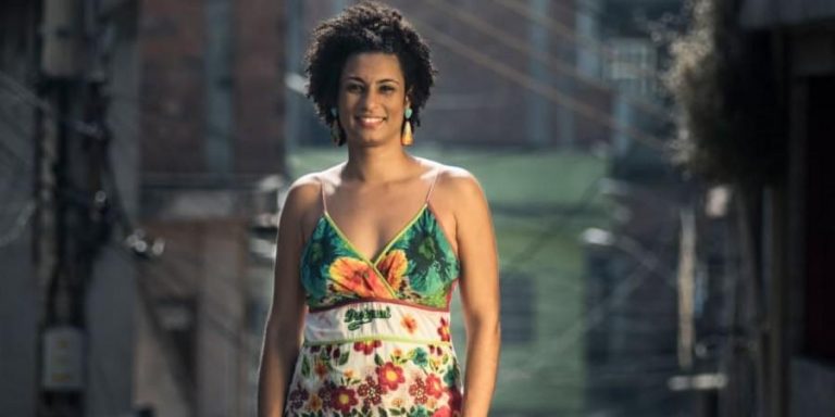 Vereadora Marielle Franco. Ela é uma mulher negra, tem o cabelo crespo e curto, usa um vestido floral e brincos coloridos. Ela olha para a câmera e sorri