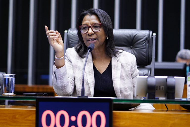 Deputada Rosangela Gomes fala ao microfone. Ela é negra, tem cabelo comprido e preto, usa óculos, uma blusa preta e um blazer claro