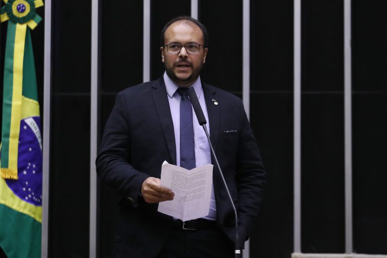 Deputado Thiago de Joaldo fala ao microfone. Ele é um homem claro, tem bigode, barba e cabelo escuro, usa um terno preto, óculos e segura um papel