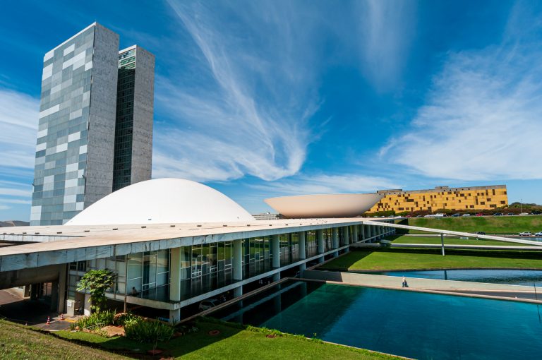 Prédio do Congresso num dia ensolarado. À frente há um espelho d'água e ao fundo um prédio comprido amarelo