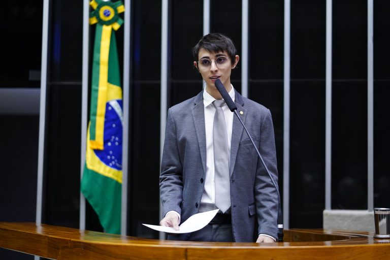 Deputado Amom Mandel fala ao microfone. Ele usa terno cinza, camisa branca e segura um papel