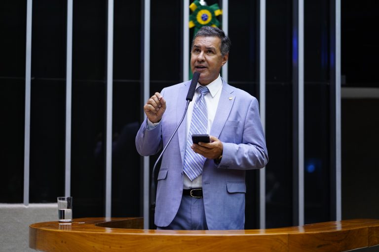Deputado Capitão Augusto fala ao microfone. Ele é um homem claro, usa terno azul claro e segura um celular
