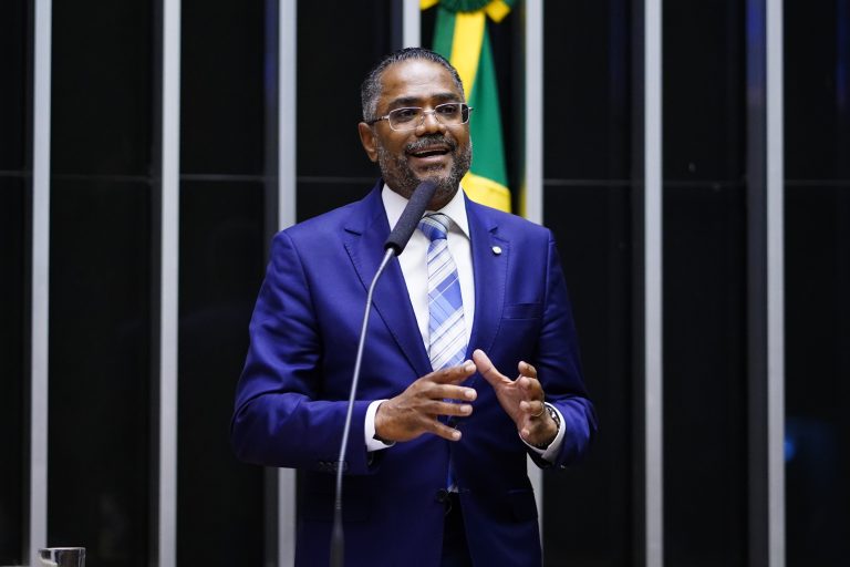 Deputado Márcio Marinho fala ao microfone. Ele é negro, tem cabelo, bigode e barba grisalhos e usa um terno azul-marinho