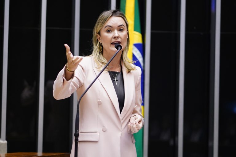 Deputada Julia Zanatta fala ao microfone. Ela é branca, tem cabelo comprido e loiro e usa um blazer bege