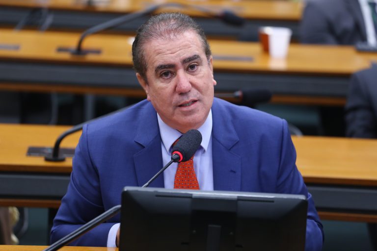Deputado Jonas Donizette fala ao microfone. Ele é um homem branco, tem o cabelo escuro e usa um terno azul, camisa branca e gravata vermelha