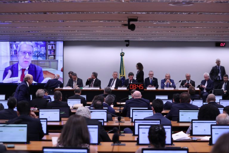 Deputados estão sentados num plenário. Ao fundo há um telão