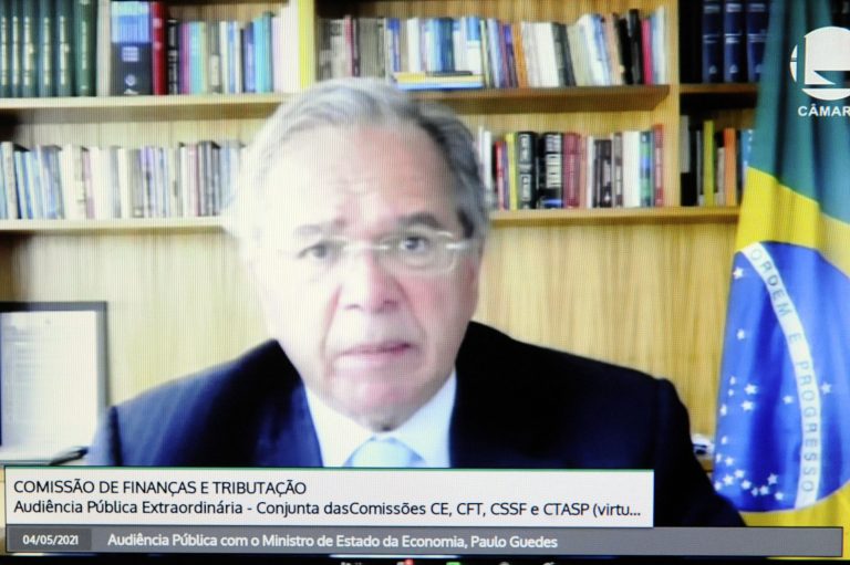 Ministro Guedes fala olhando para a tela do computador. Atrás dele há uma estante de livros e uma bandeira do Brasil