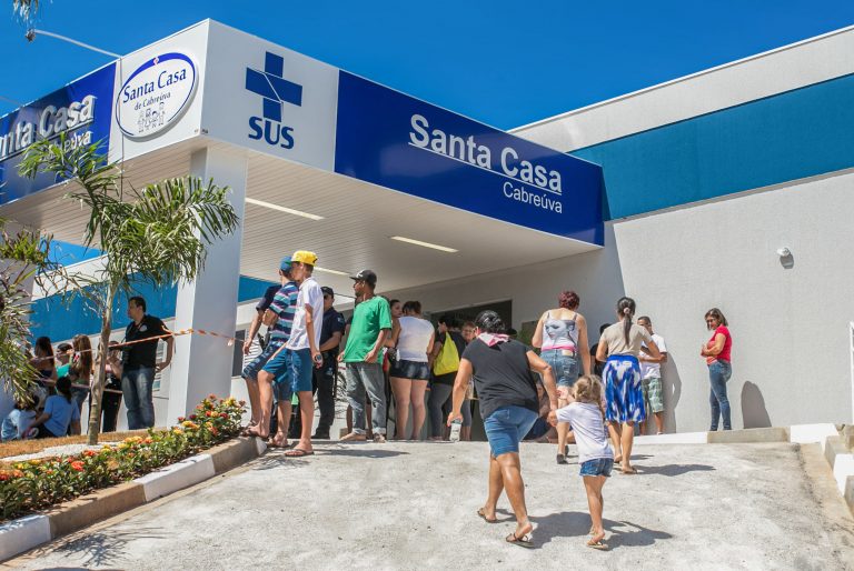 Fachada de um prédio branco e azul onde se lê: Santa Casa. Na porta há várias pessoas em pé aguardando atendimento