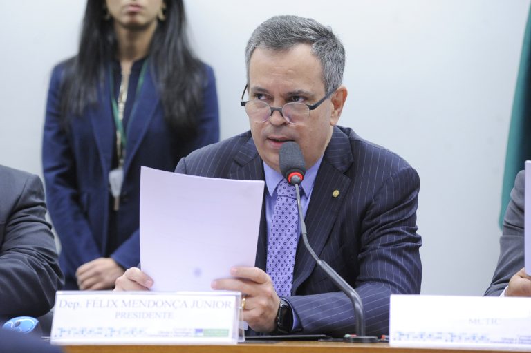 Deputado Félix Mendonça Júnior está sentado falando ao microfone. Ele usa um terno escuro, usa óculos e segura um papel