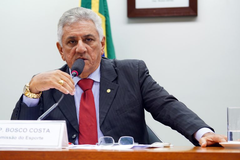 Deputado Bosco Costa está sentado falando ao microfone