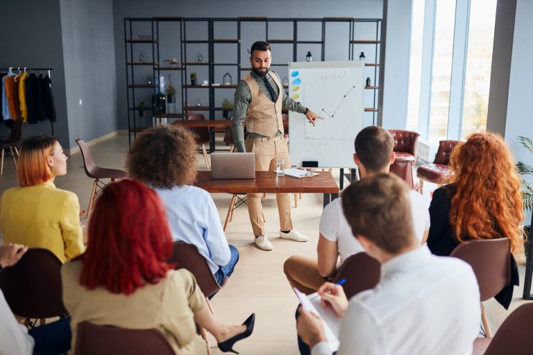 Grupo de pessoas está reunido ouvindo um homem falar. Ele está em pé mostrando um gráfico