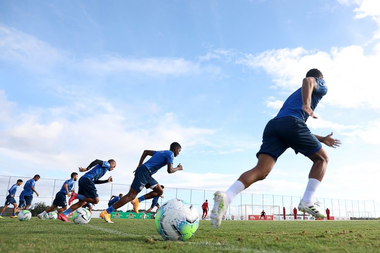 Jovens treinam num campo de futebol. Eles usam unifromes azuis e correm num gramado. Ao fundo, o céu claro.