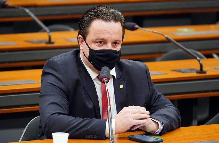 Deputado Rodrigo Coelho está sentado falando ao microfone. Ele usa máscara facial e veste um terno escuro