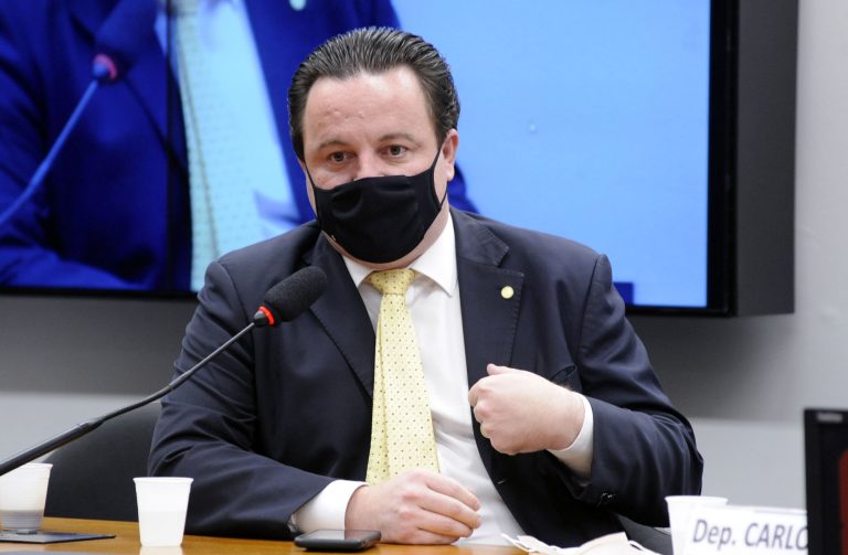 Deputado Rodrigo Coelho está sentado falando ao microfone. Ele usa máscara facial e veste um terno escuro