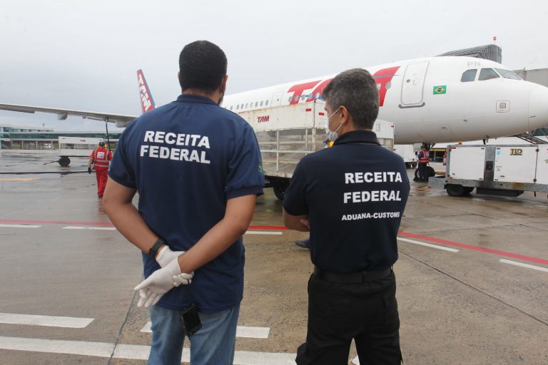 Dois agentes da Receita Federal estão em pé, de costas, numa pista de pouso. Ao fundo há um avião estacionado