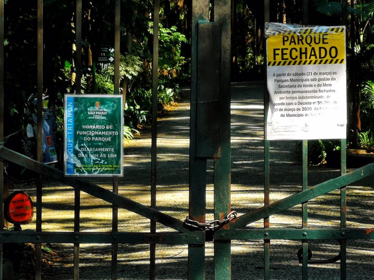 Portão de parque público fechado por causa da pandemia. Na grade verde está afixado um cartaz onde se lê: "Parque fechado"