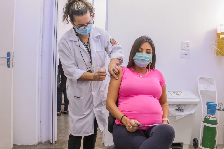 Mulher grávida toma vacina contra a Covid-19. Ela usa uma blusa rosa e está sentada enquanto a enfermeira aplica a vacina