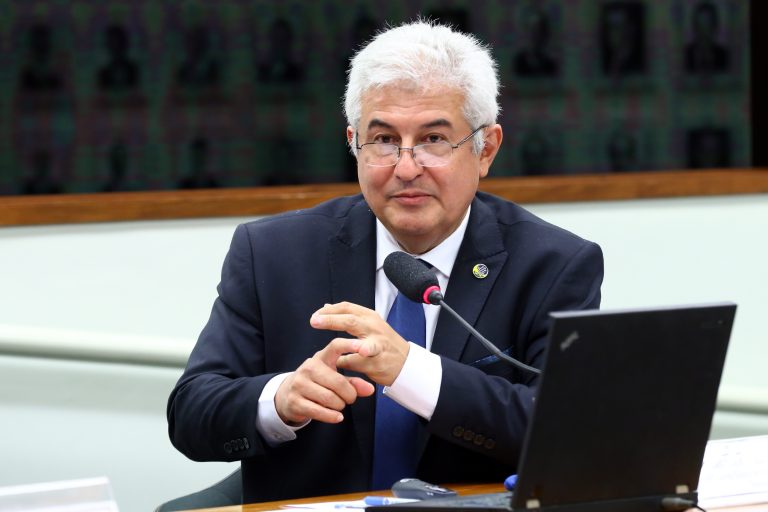 Ministro de Ciência e Tecnologia, Marcos Pontes, está sentado falando ao microfone. Ele tem cabelo branco, usa óculos e veste um terno escuro