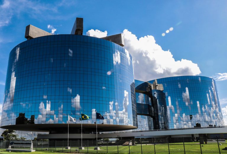 Fachada de um prédio curvo todo espelhado. Ao fundo o céu azul