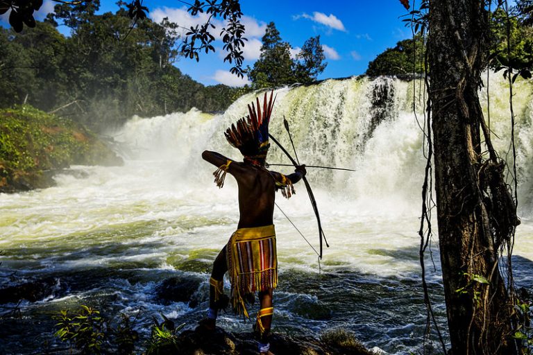 Um índio usa arco e flecha para pescar um rio. Ao fundo, uma cachoeira e um céu azul