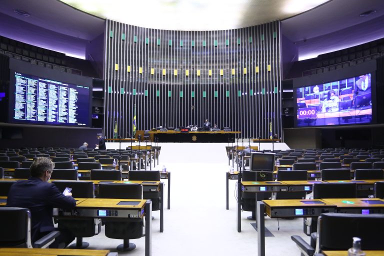 Plenário da Câmara quase vazio com um telão ao fundo por meio do qual os deputados participam remotamente das votações