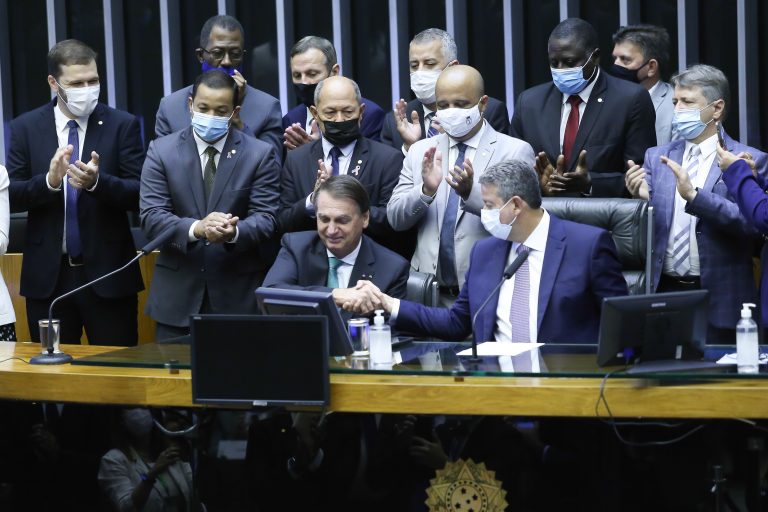 Presidente Bolsonaro está sentado e aperta a mão do presidente da Câmara que está sentado ao lado