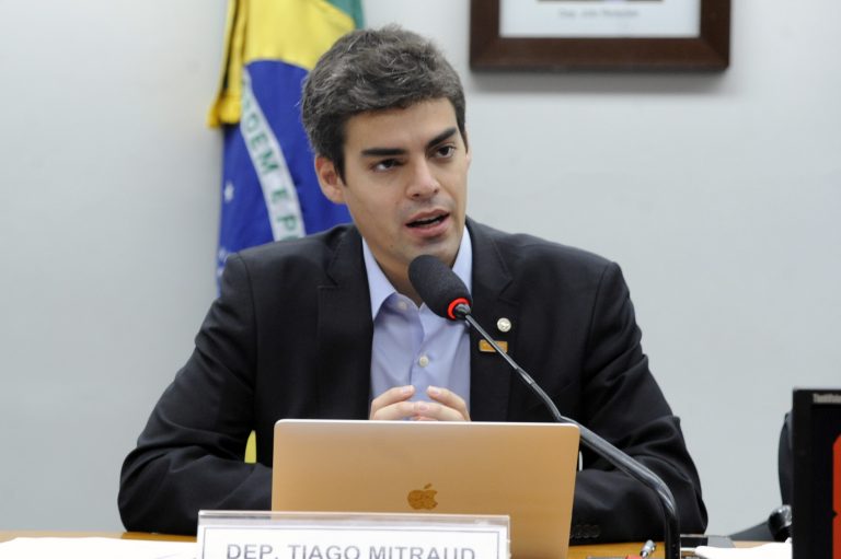 Deputado Tiago Mitraud está sentado falando ao microfone. Ele tem cabelo escuro e pele clara e veste um terno preto