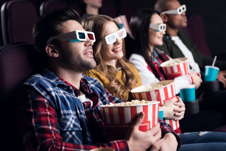 Pessoas estão sentadas num cinema usando óculos três D e segurando baldes de pipoca