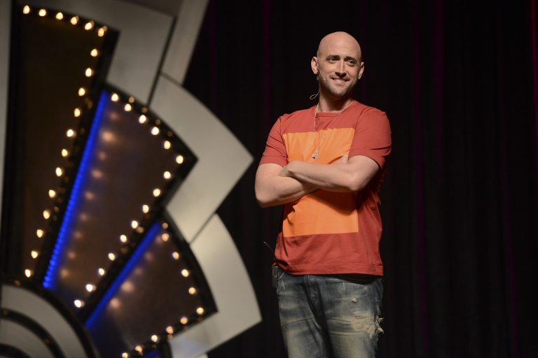 Um ator está em pé num palco. Ele veste uma camisa laranja e é careca. Ao fundo há um pano preto e luzes do lado esquerdo