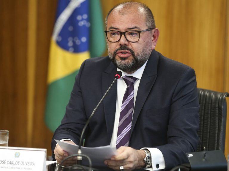 Ministro do Trabalho fala ao microfone. Ele é um homem branco, meio calvo, usa óculos e um terno escuro e segura um papel. Ao fundo, do lado esquerdo tem uma bandeira do Brasil