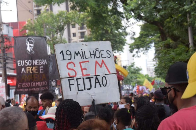 Pessoas protestam contra a fome. Elas estão reunidas numa rua e seguram cartazes