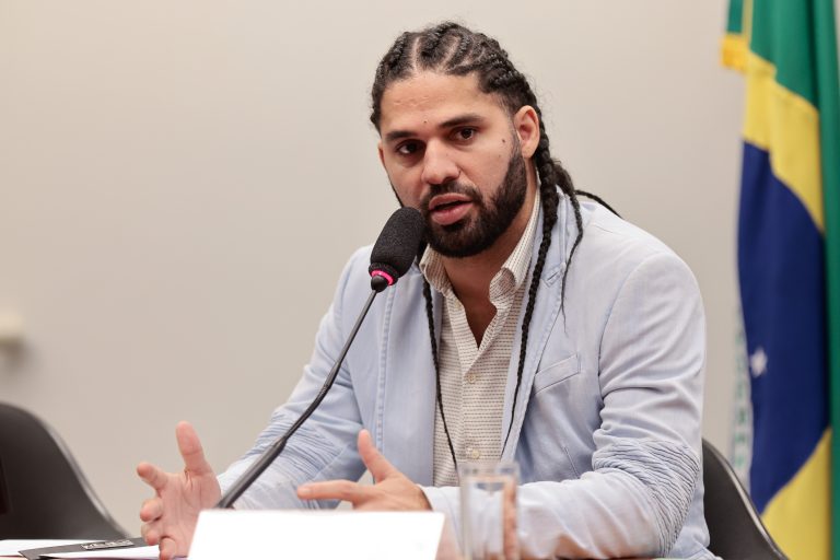 Deputado David Miranda fala ao microfone. Ele usa barba e bigode e tem o cabelo longo com tranças