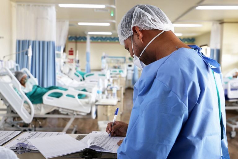 Um enfermeiro veste uniforme azul e touca e máscara brancas e faz anotações num papel. Ao fundo há vários pacientes deitados