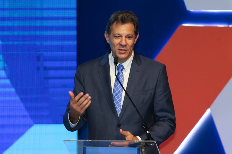 O ministro da Fazenda fala ao microfone. Ele é um homem branco, com cabelo escuro e veste um terno preto com camisa branca