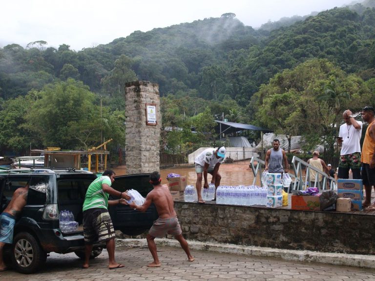 Homens carregam garrafas de água num carro. Ao fundo, há um rio barroso e uma encosta com mata