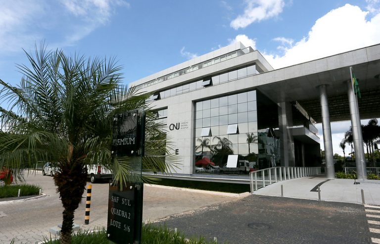 Foto de um prédio baixo com fachada de vidro num dia ensolarado e as letras CNJ na fachada, No primeiro plano aparece uma palmeira