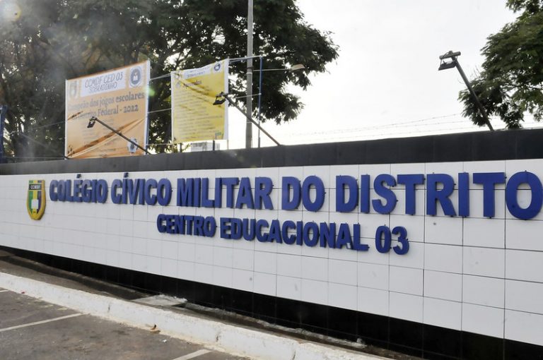 Fachada de uma escola. Um muro branco com o nome da escola escrito em letras azuis