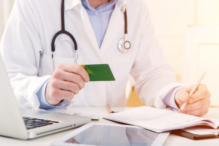 Um médico segura um cartão em uma mão enquanto escreve num papel. A foro está cortada e não é possível ver o rosto dele
