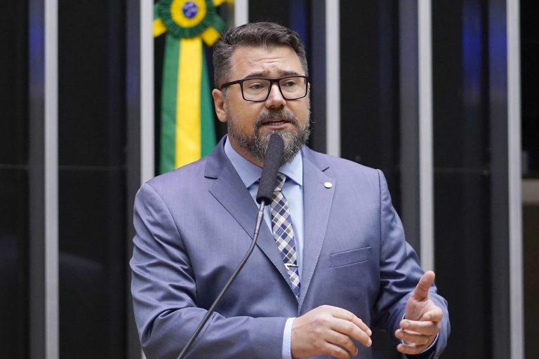 Deputado Marcos Pollon fala ao microfone. Ele é um homem branco com barba e bigode, usa óculos e terno cinza