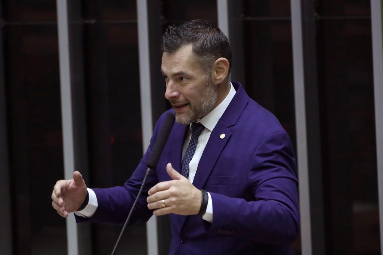 Deputado Marangoni fala ao microfone. Ele é um homem branco, com bigode e barba grisalhos e usa um terno azul-marinho
