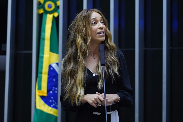 Deputada Erika Hilton fala ao microfone. Ela tem o cabelo longo e loiro e veste um vestido preto
