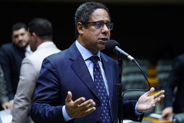 Deputado Orlando Silva fala ao microfone. Ele é negro, usa terno escuro e camisa clara