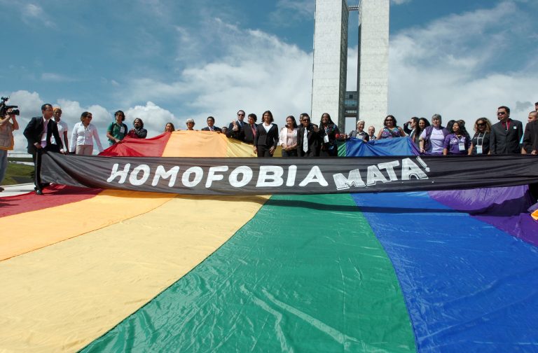 Pessoas seguram em frente ao Congresso Nacional uma bandeira com listras coloridas e uma faixa escrito: Homofobia mata