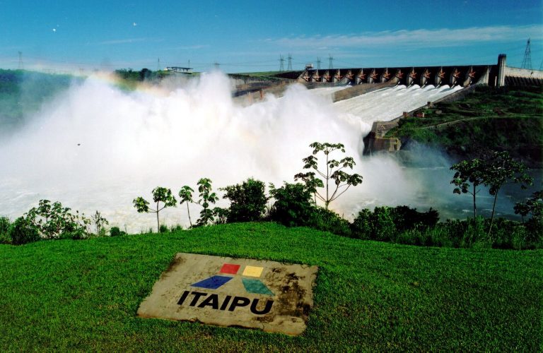 Foto da hidrelétrica de Itaipu. Ao fundo há uma represa de onde cai a água. À frente há árvores, um gramado e uma placa escrito Itaipu