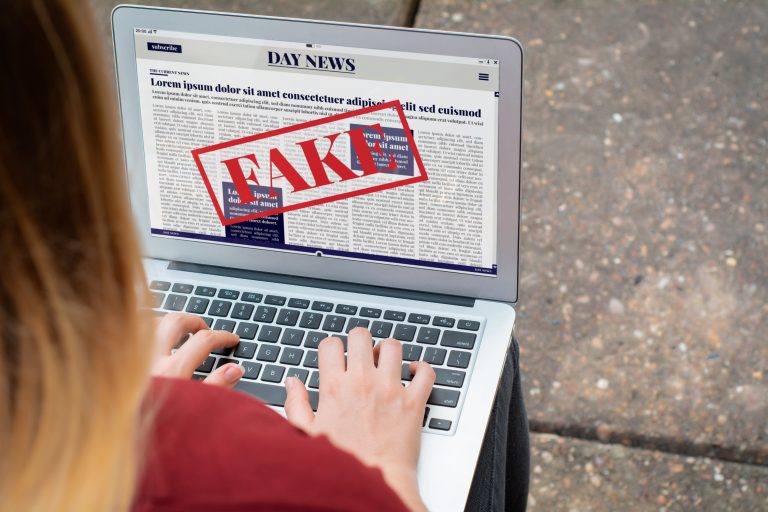 Uma mulher mexe no laptop. Na tela está escrito "Fake" em cima de um jornal