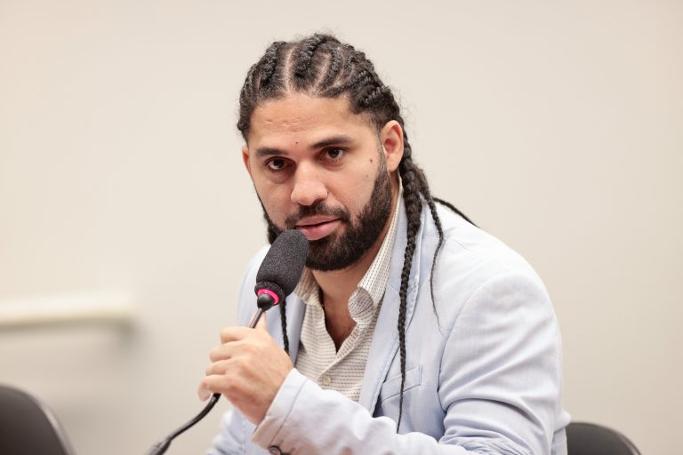 Deputado David Miranda fala ao microfone. Ele usa barba e bigode e tem o cabelo longo com tranças