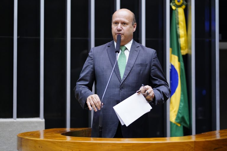 Deputado Célio Silveira fala ao microfone. Ele é um homem branco, calvo, usa um terno cinza e gravata verde, e segura um papel